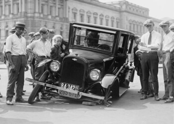 A vintage car crash attended by a group of onlookers in a 1920s urban setting.